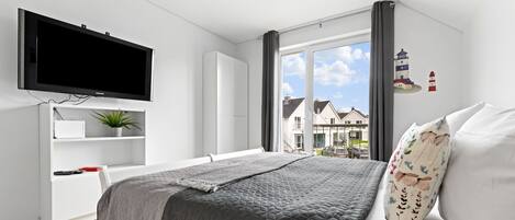 Une chambre moderne avec un grand lit, un téléviseur à écran plat, un bureau agrémenté d’une plante et une vue sur les maisons par la fenêtre.