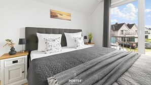 A modern bedroom with a large bed, a nightstand, a window with a view of houses, and a painting on the wall. - Ocean Time - 1st Floor, 2 Balconies, Infrared Sauna, Fireplace, Sun Loungers (Olpenitz)