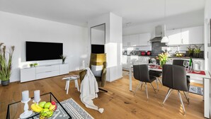 A modern living room with a flat-screen TV, a dining area with black chairs, and a kitchen with white cabinets and a black stove. - Ocean View 14 - Balcony, Grill, Fireplace, Shower, Elevator, Underground Parking (Olpenitz)