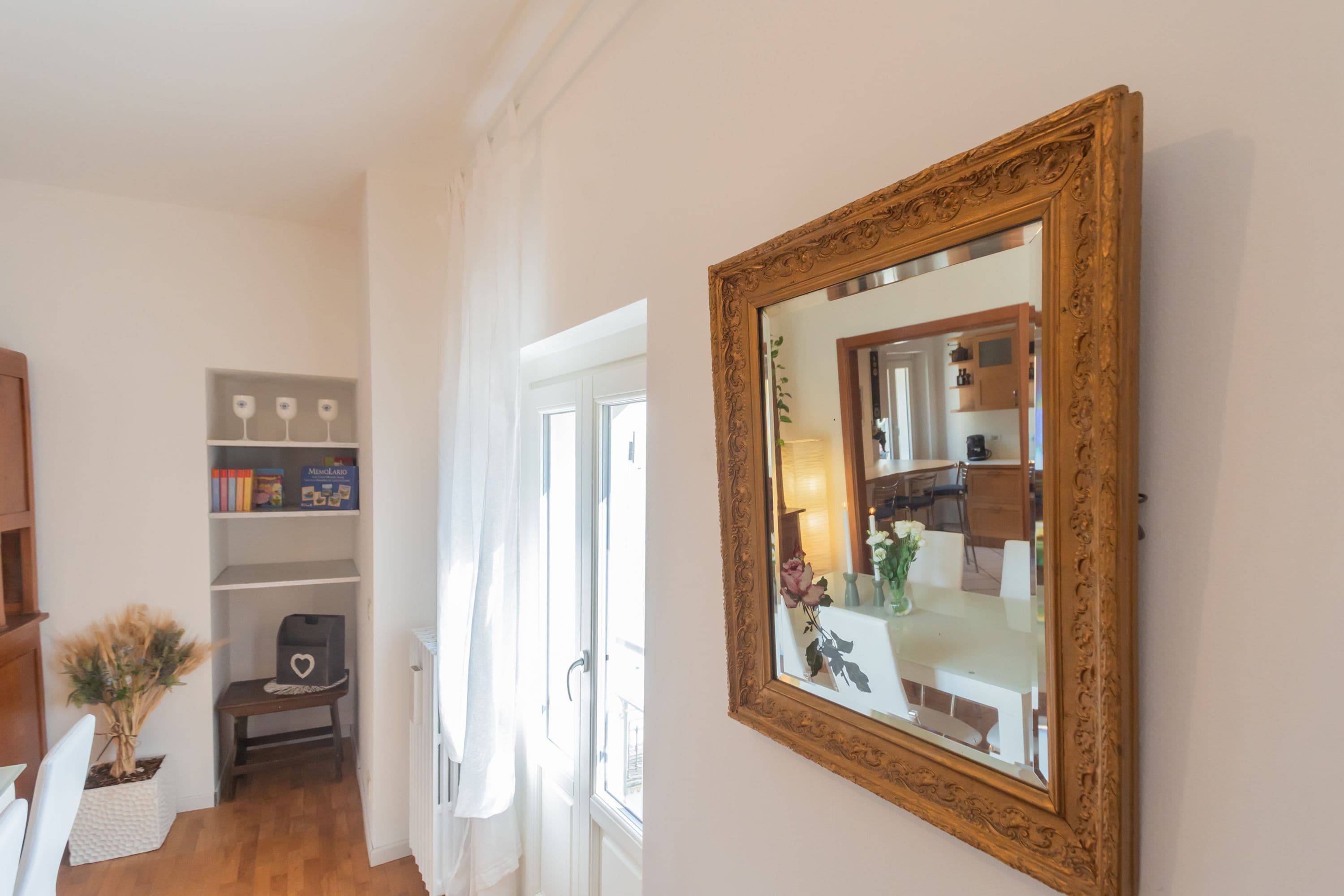 A hallway with a wooden floor, white walls, and a large ornate mirror. There is a bookshelf with books and decorative items, and a small table with a vase of flowers.