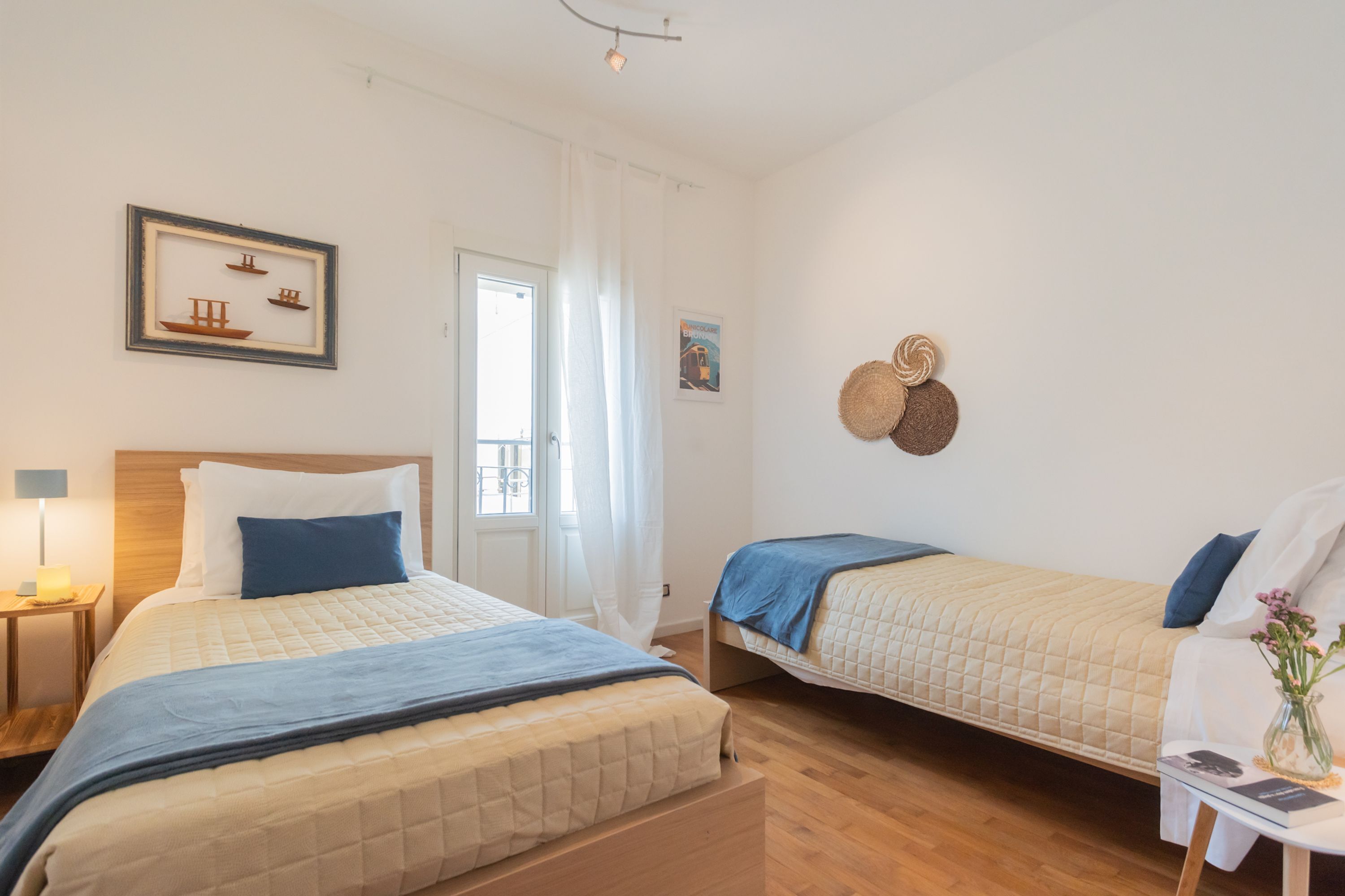 Een hotelkamer met twee bedden, een houten vloer, een ingelijste foto aan de muur, een raam met gordijnen en een klein tafeltje met een lamp en een bloemenvaas.