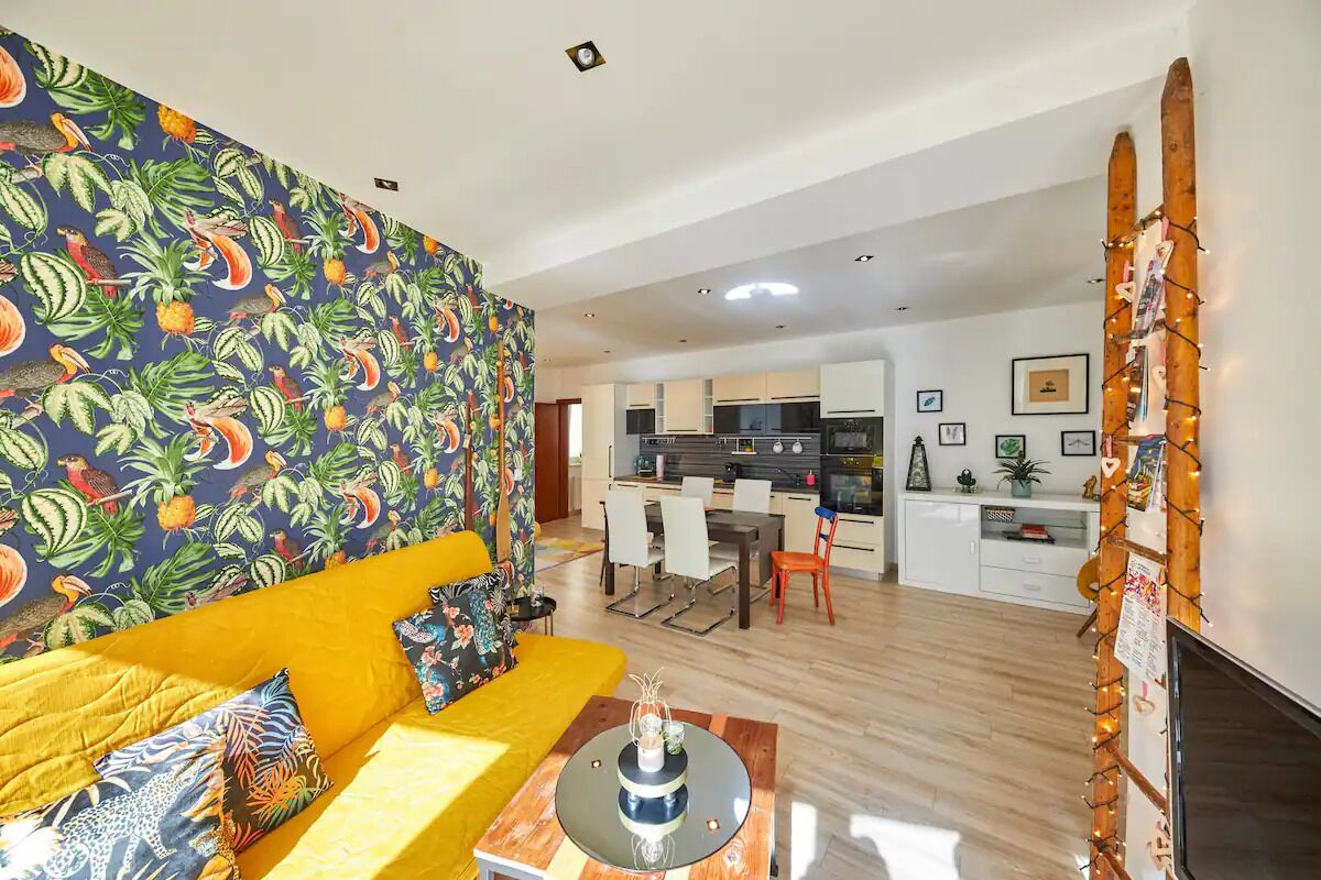 A modern living room with a vibrant yellow sofa, a patterned wall, and a dining area with a kitchen in the background.