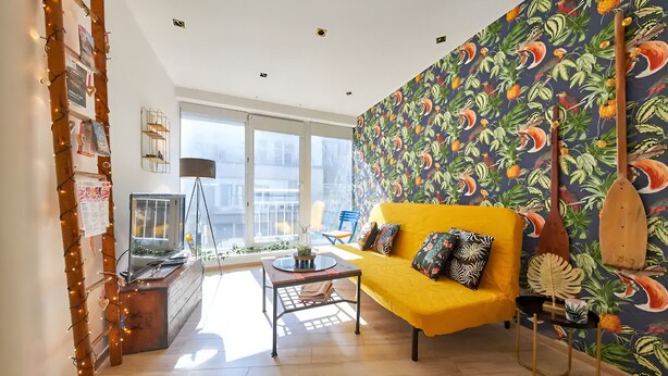 Un salon lumineux avec un canapé jaune, un mur orné de motifs et une vue sur la ville grâce à de grandes fenêtres.