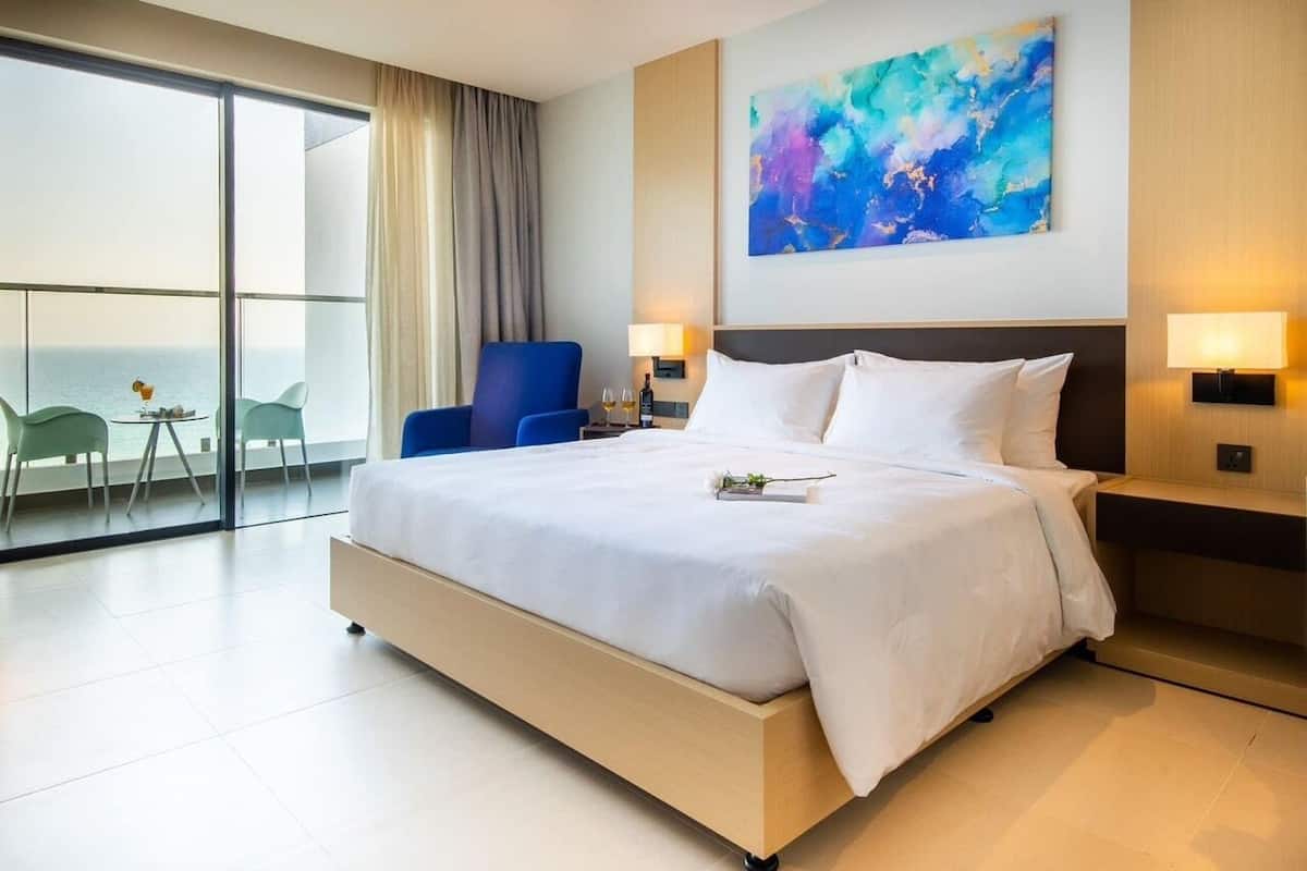 Una habitació d'hotel moderna amb un llit gran, vista al mar, una cadira blava i un quadre a la paret.