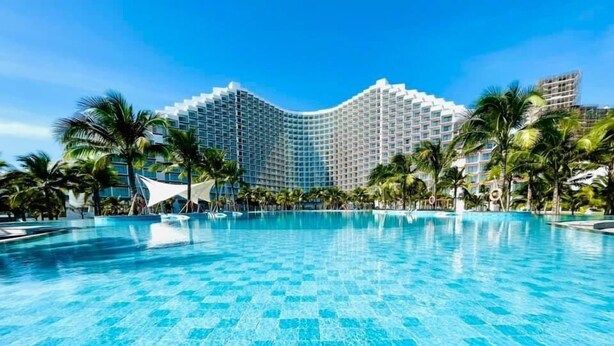 A large, clear swimming pool with a tiled floor, surrounded by palm trees and a modern hotel building in the background. - The Sea View Cam Ranh (Bac Cam Ranh)