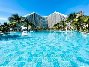 A large, clear swimming pool with a tiled floor, surrounded by palm trees and a modern hotel building in the background. - The Sea View Cam Ranh (Bac Cam Ranh)