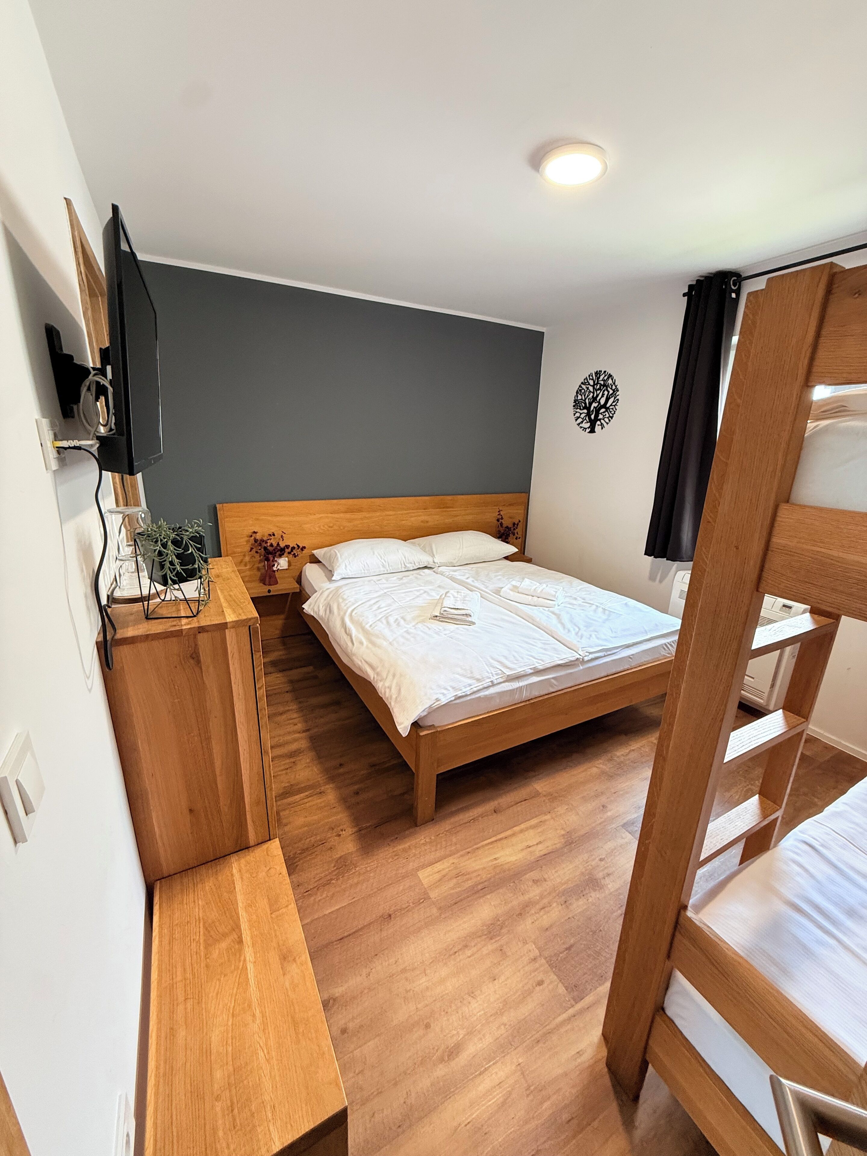 A small, cozy room with a single bed, a wooden nightstand, a TV mounted on the wall, and a ceiling light.