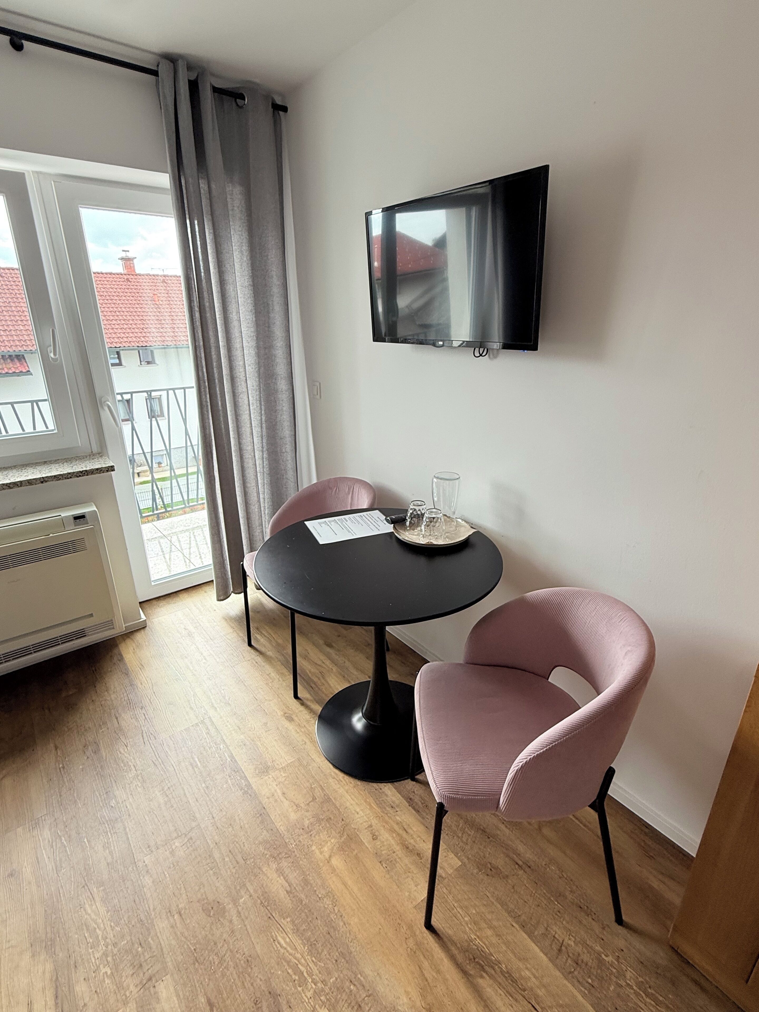 Una habitación moderna con una mesa redonda negra, dos sillas rosas, un televisor de pantalla plana montado en la pared y una ventana con vistas a edificios.