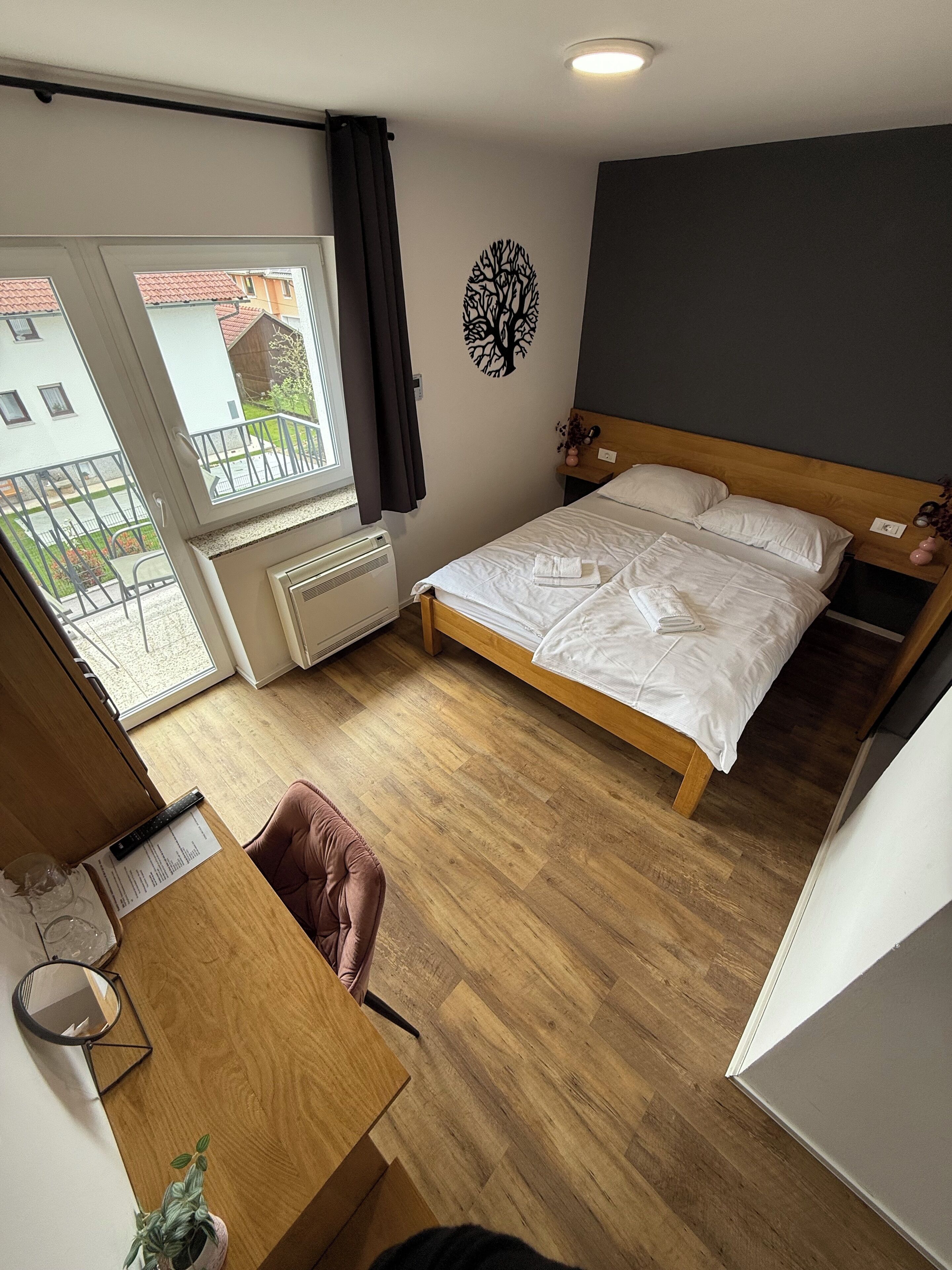 A bedroom with a wooden bed, a desk with a chair, a radiator, and a window with a view of buildings.