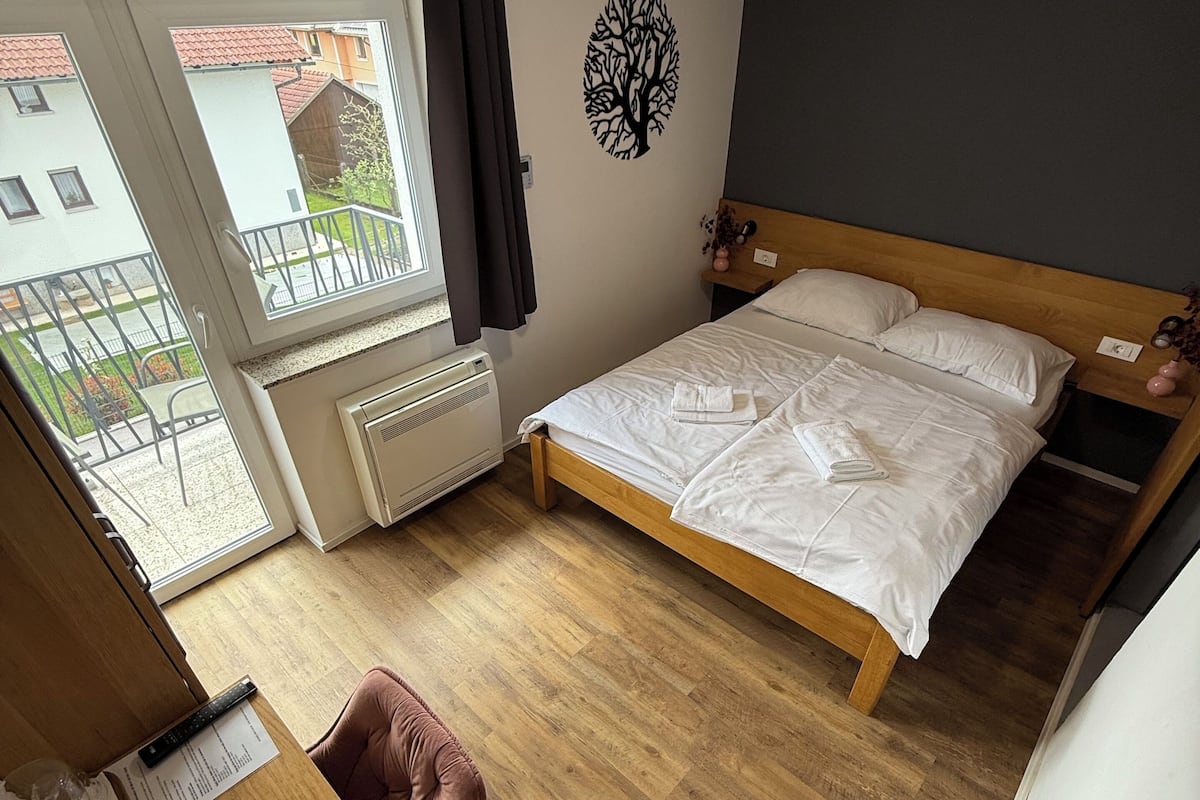 A bedroom with a wooden bed, a desk with a chair, a radiator, and a window with a view of buildings.