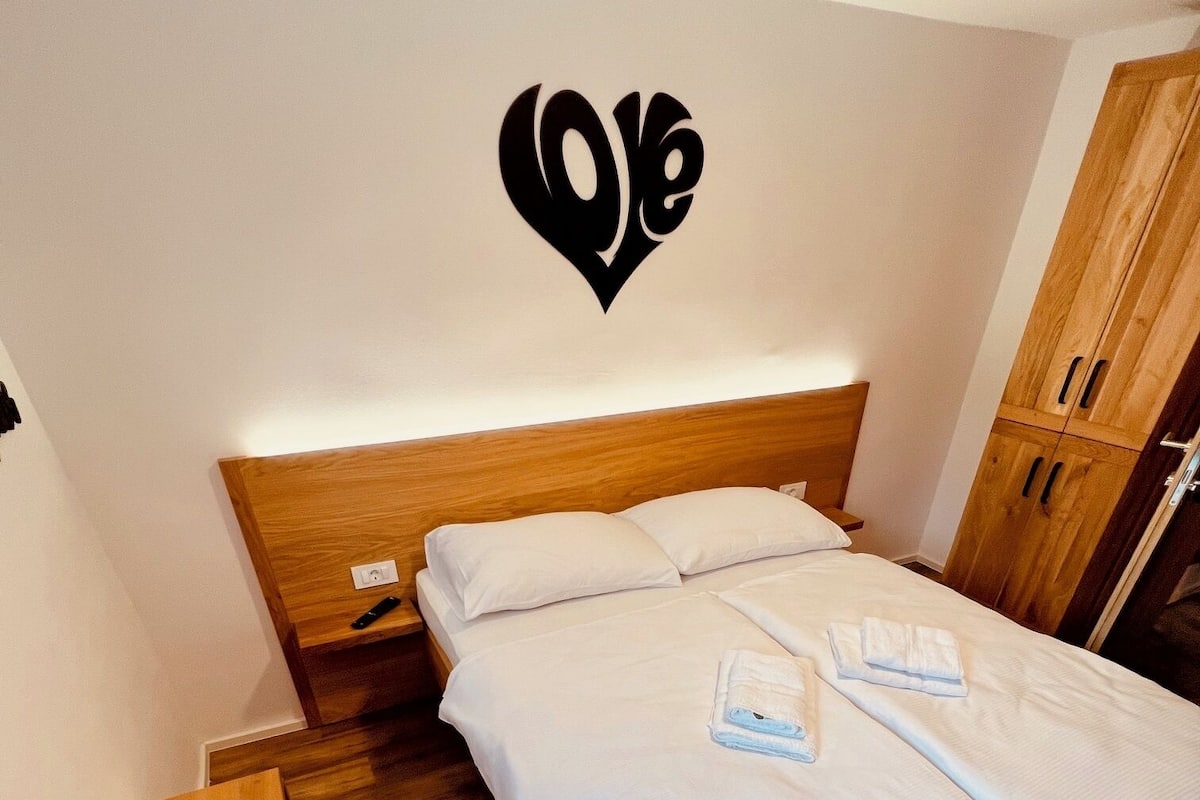 A neatly made bed with white linens and two folded towels, a wooden headboard, a wooden wardrobe, and a wall-mounted heart-shaped decoration.