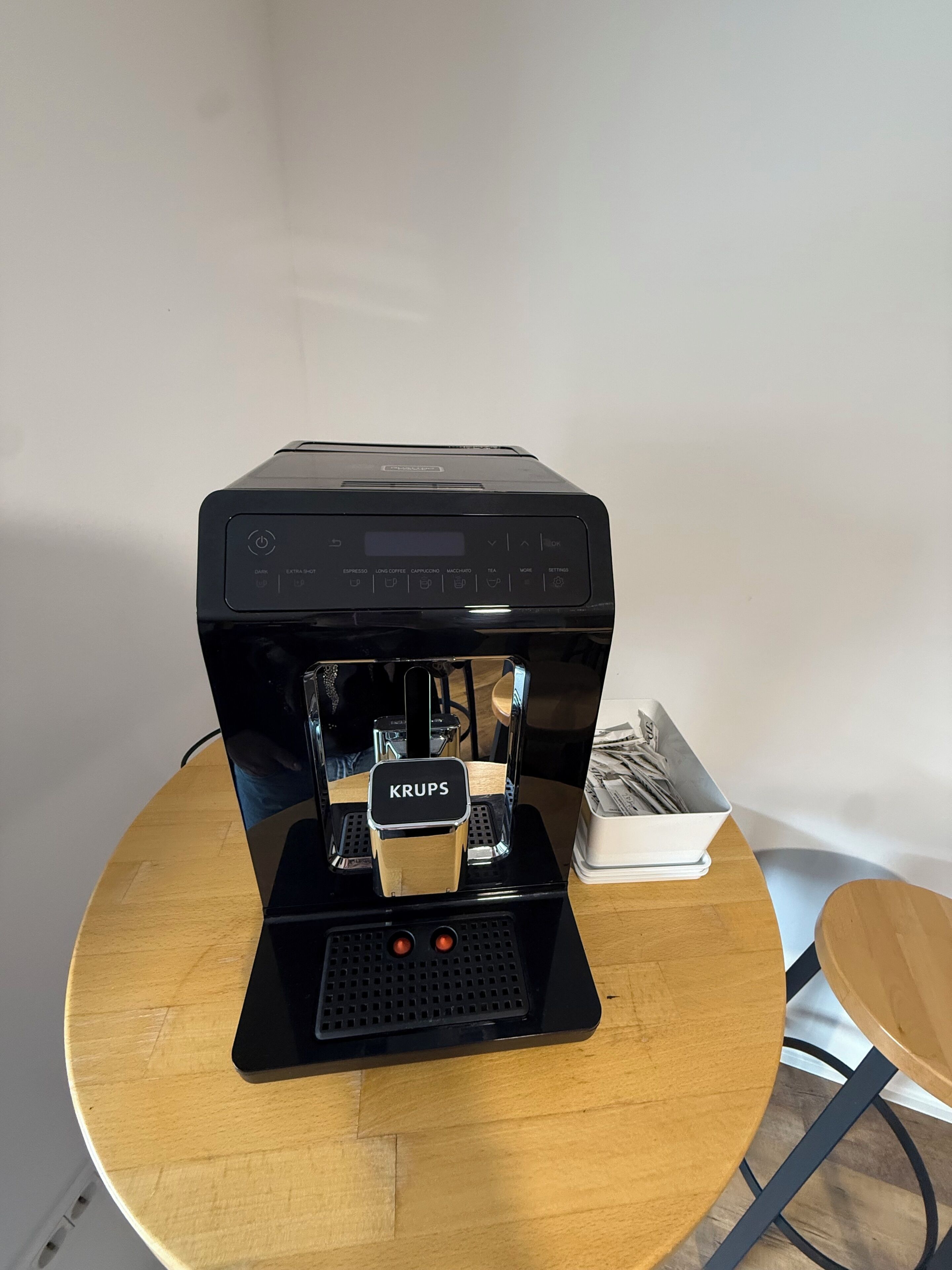 A Krups coffee machine on a wooden table with a white cup holder nearby.