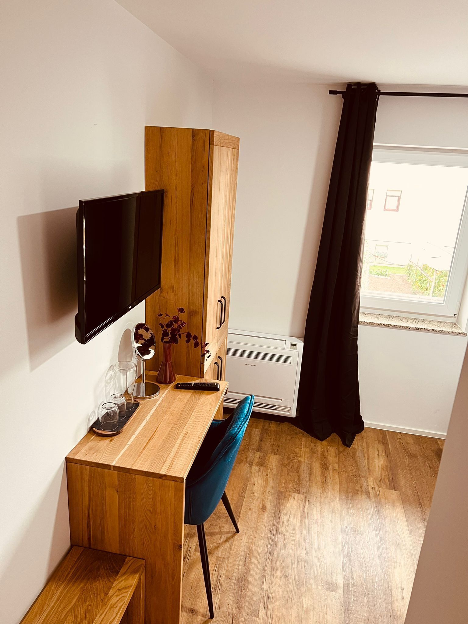 Ein kompakter Raum mit einem Holztisch, einem blauen Stuhl, einem Wand-Fernseher, einem kleinen Schrank und einem Fenster mit schwarzem Vorhang.