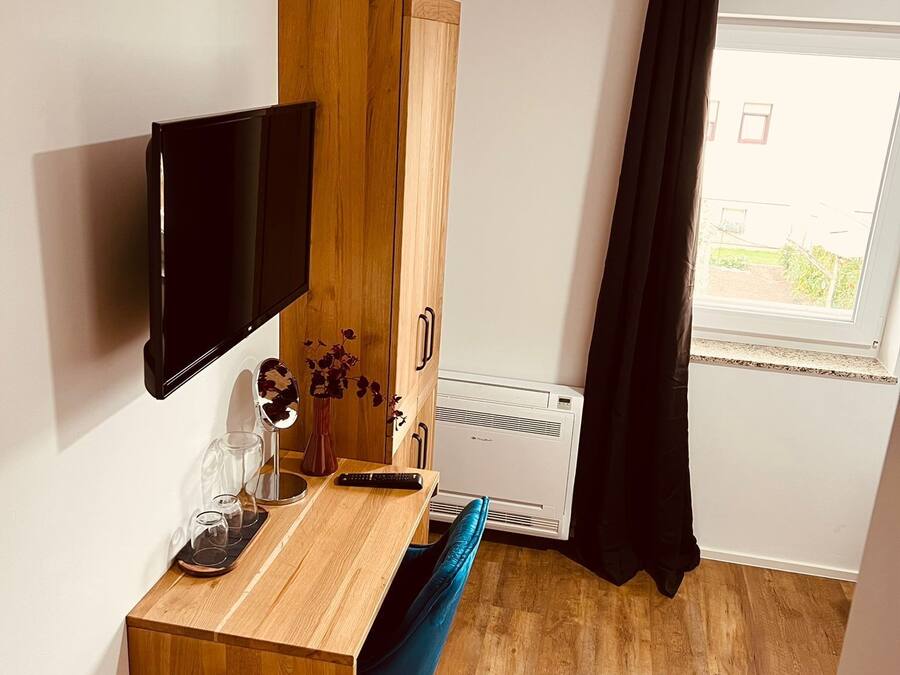Ein kompakter Raum mit einem Holztisch, einem blauen Stuhl, einem Wand-Fernseher, einem kleinen Schrank und einem Fenster mit schwarzem Vorhang.