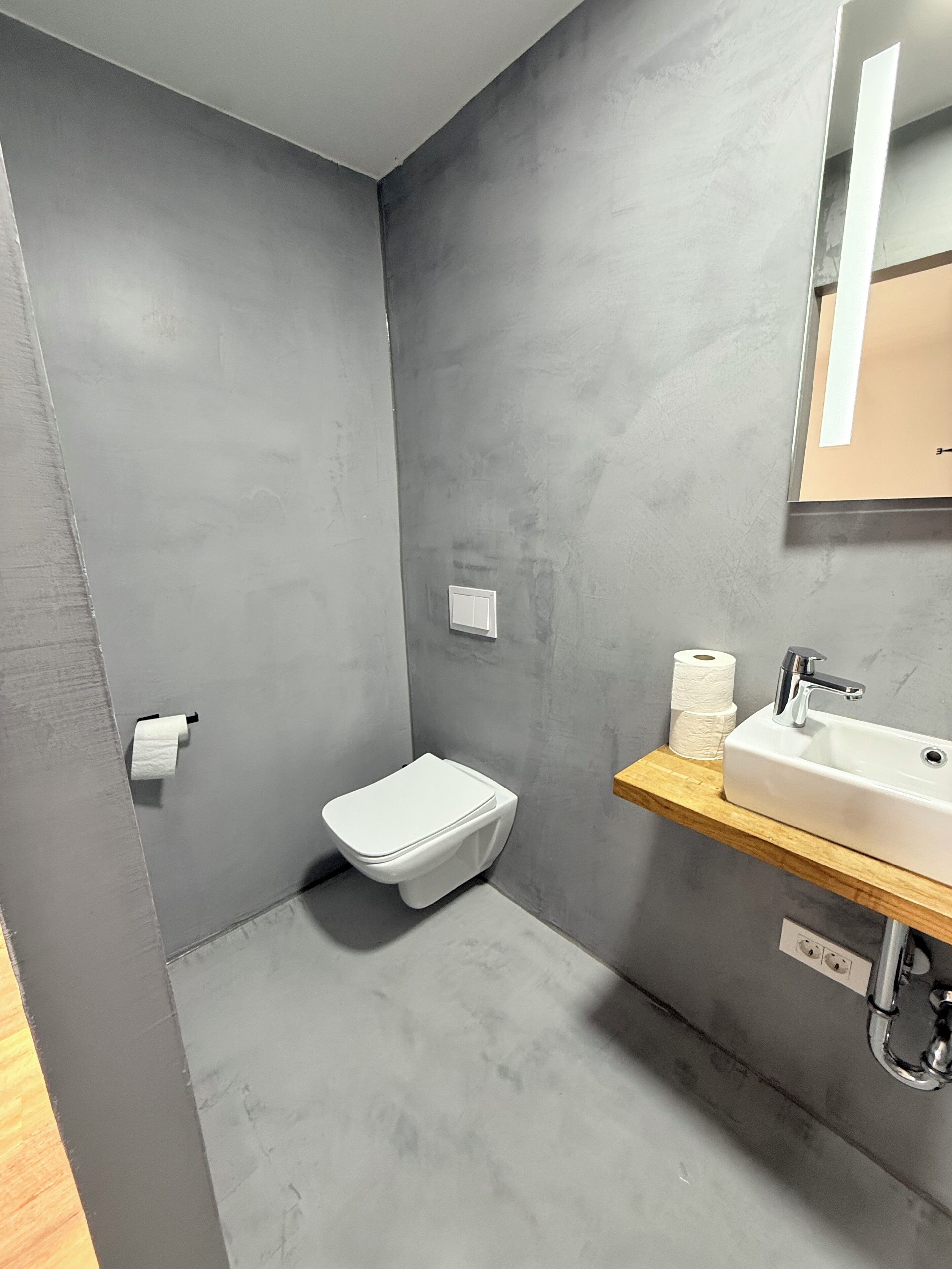 A modern bathroom with a grey tiled wall, a white toilet, a wooden shelf with a sink, and a mirror.