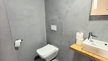 A modern bathroom with a grey tiled wall, a white toilet, a wooden shelf with a sink, and a mirror.