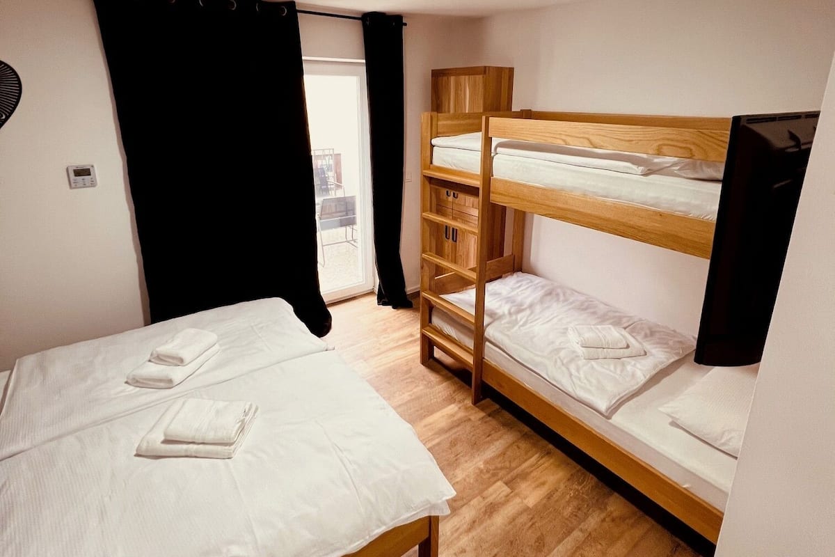 A room with a single bed, a bunk bed, wooden flooring, a window with black curtains, and a ceiling light.