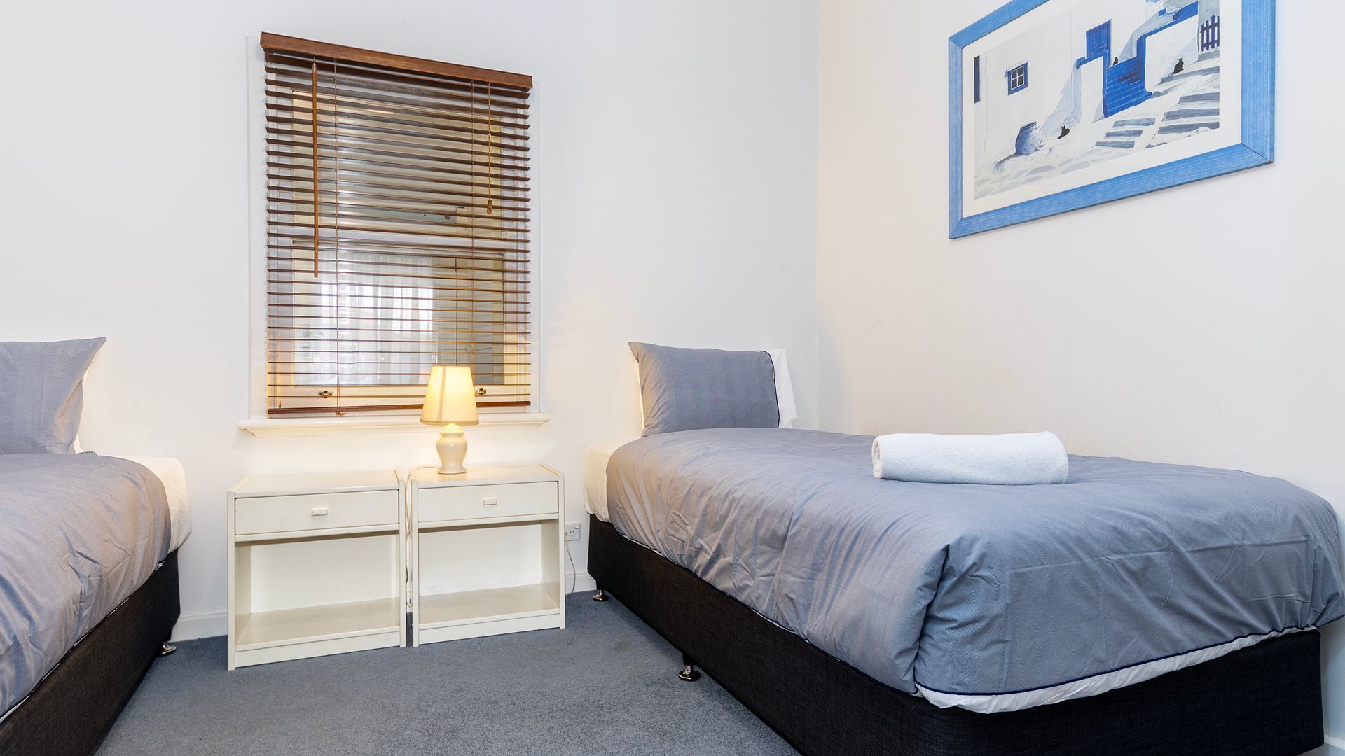 Een slaapkamer met twee bedden, een nachtkastje met een lamp, een klein wit kastje en een lijst met een foto aan de muur.