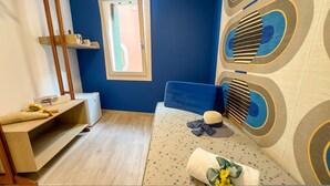 A room with a blue wall, a window, a bed with a patterned headboard, a bedside table with a lamp, and a wall-mounted shelf with a cup and a book. - Ca de la Corona - Ipnosi by Ca-sa Check (Chioggia)