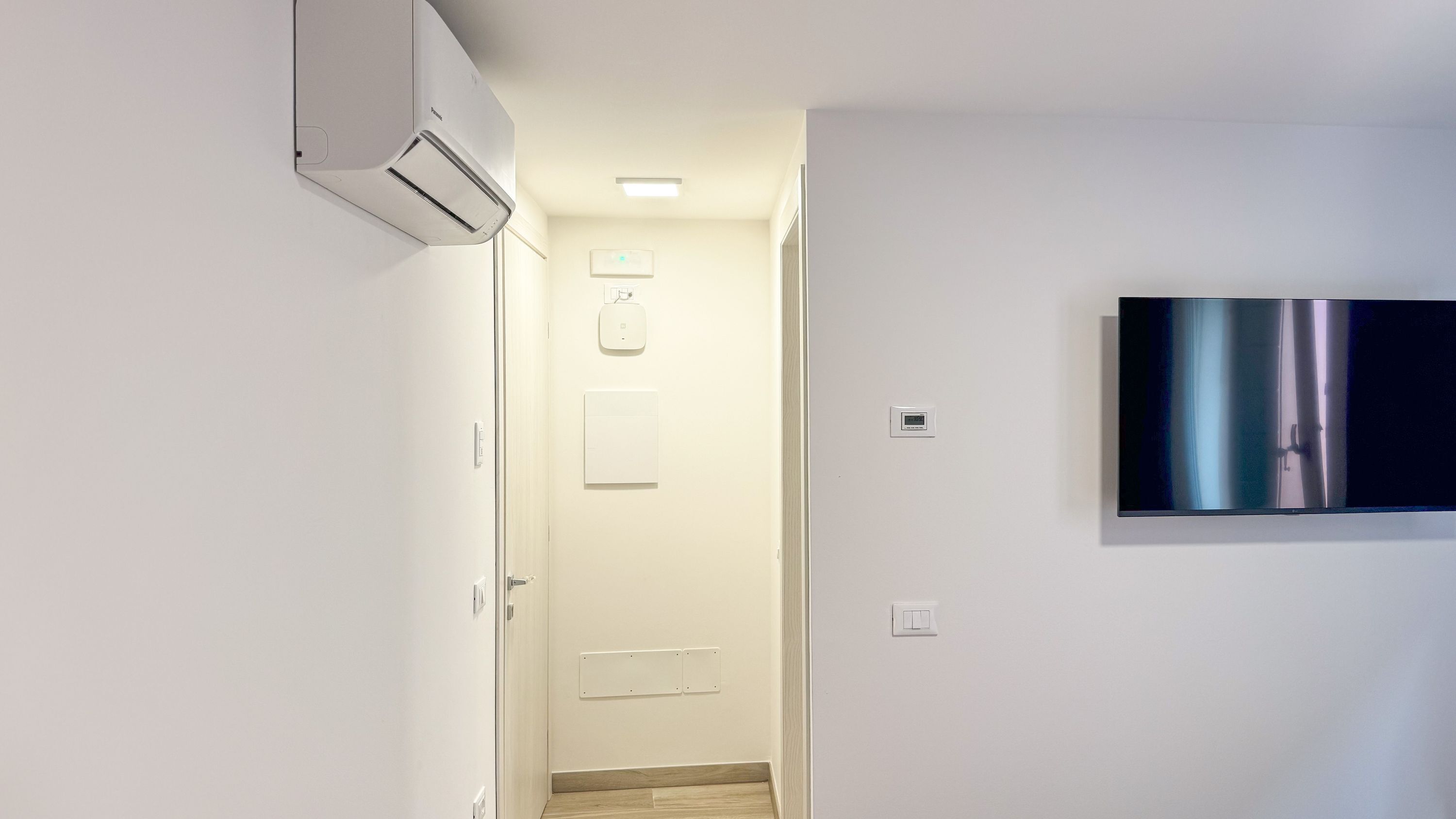 Une chambre d’hôtel moderne équipée d’un téléviseur à écran plat fixé au mur, d’un système de climatisation intégré, d’une porte avec une poignée et d’un interrupteur.