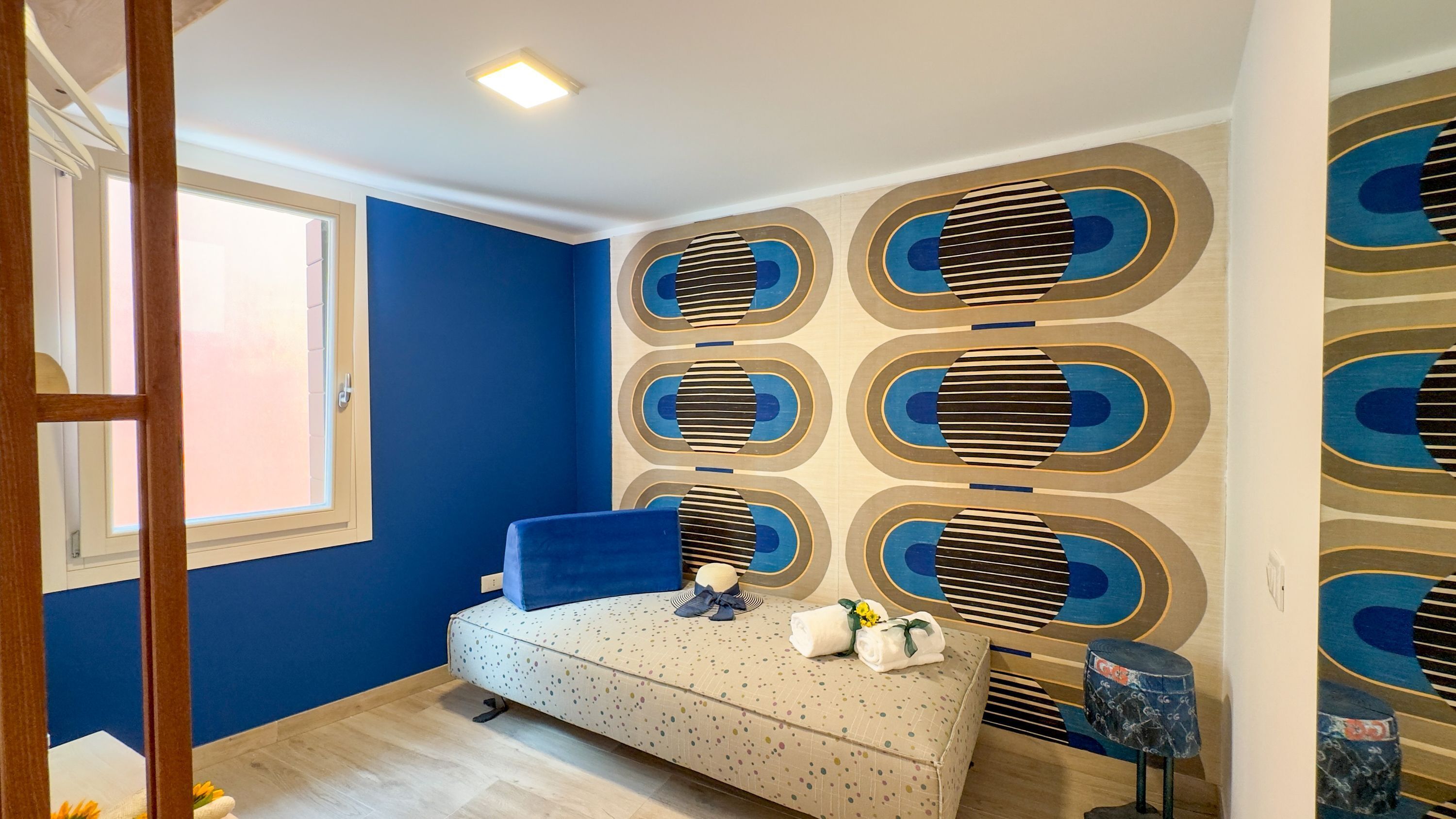 Une chambre à coucher avec un lit, une chaise bleue, un mur orné d’un motif géométrique et une fenêtre.