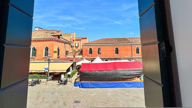 Une vue à travers une porte ouverte, donnant sur un bâtiment doté d’un toit en tuiles et d’une façade rouge.