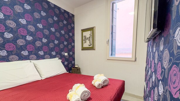 A hotel room with a red bedspread, two rolled towels, a clock on a bedside table, and a window with a view.