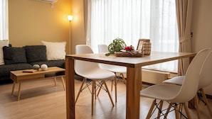 A modern living room with a wooden dining table, white chairs, a sofa with cushions, a small side table, and a potted plant. - Lilac (Sapporo)