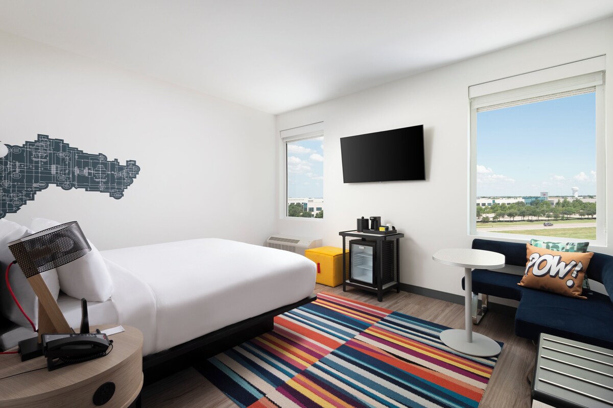 A hotel room with a bed, a desk, a chair, a TV, a window with a cityscape view, and a colorful striped rug.
