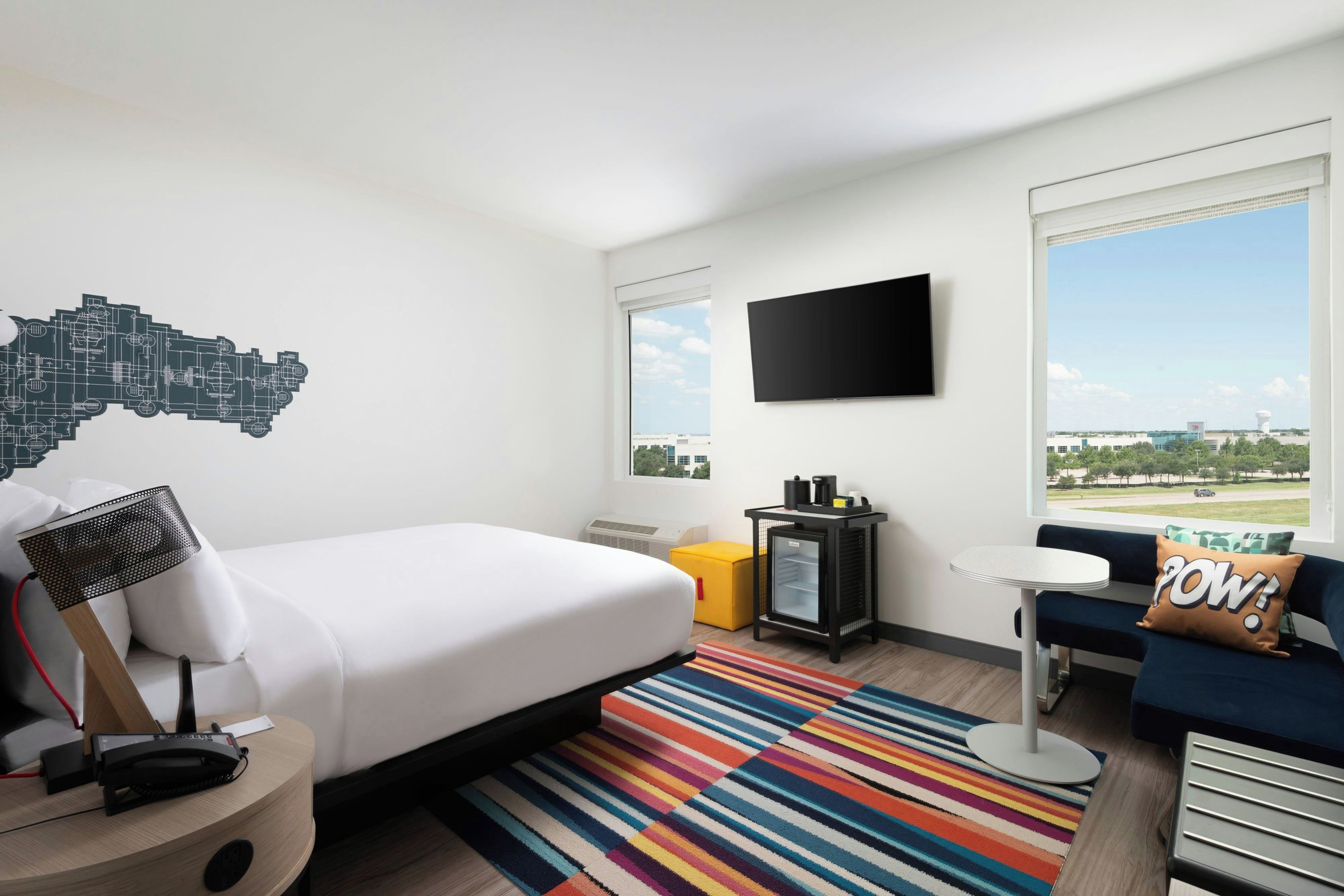 A hotel room with a bed, a desk, a chair, a TV, and a window with a cityscape view.