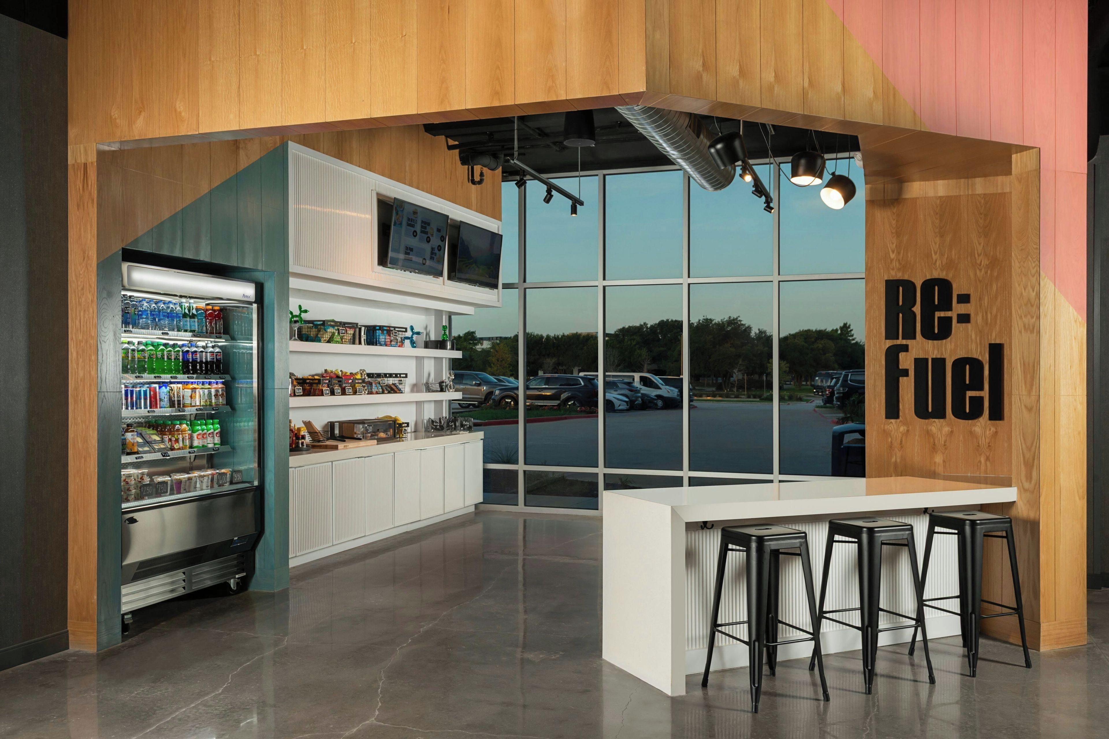 A modern bar area with a large window, a refrigerator stocked with beverages, and a counter with stools.