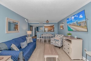 A living room with a blue sofa, a dining area with a table and chairs, a kitchen with wooden cabinets, and a large TV displaying a beach scene.