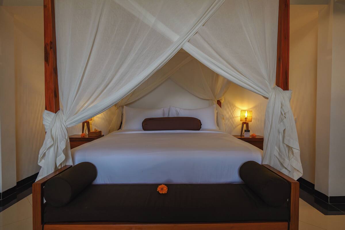 A bed with a canopy, a nightstand with a lamp, and a wooden bench with a flower.