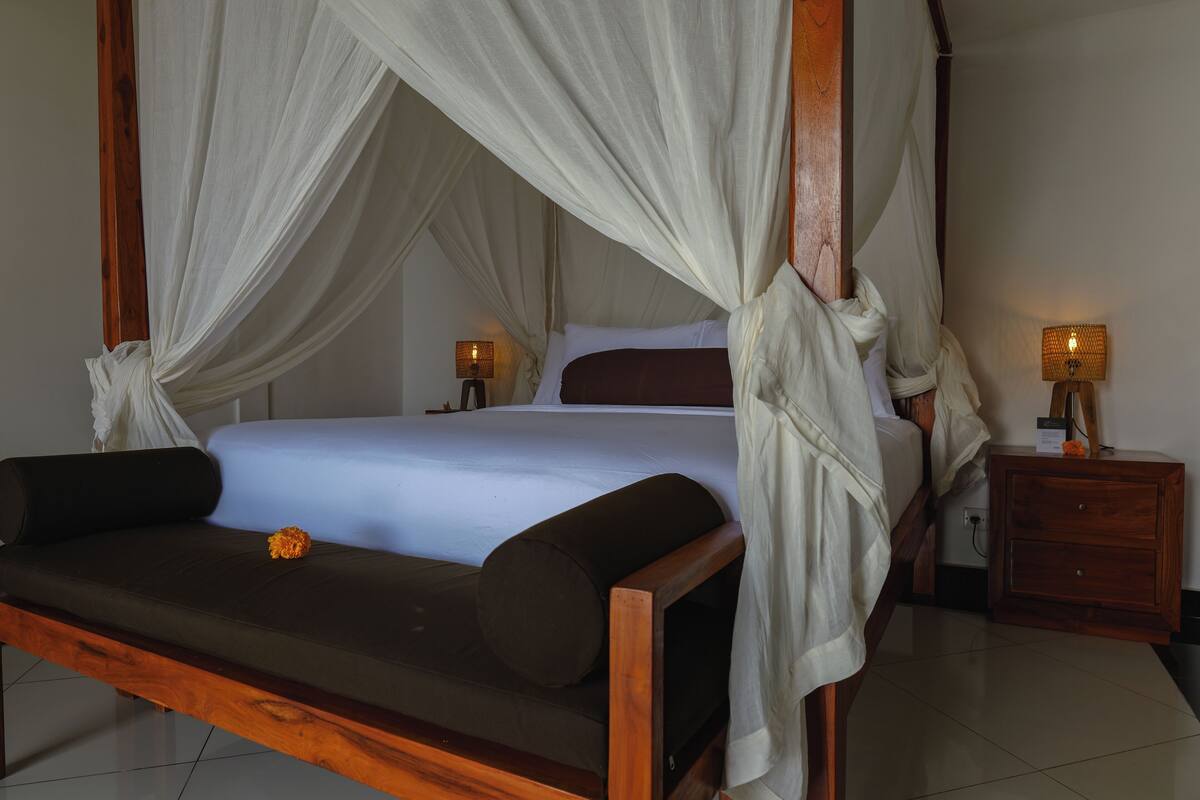 A four-poster canopy bed with a wooden frame, white bedding, and a single orange flower on the bed. There is a wooden nightstand with a lamp beside the bed.