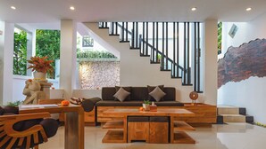 A modern living room with a sofa, coffee table, and wooden chairs. A staircase with black railings leads to an upper level. - Elemara  (Ubud)