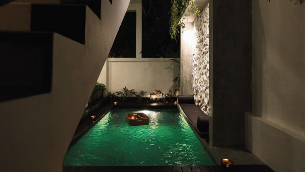 A nighttime swimming pool with a floating platform, surrounded by a modern architectural design with a stone wall and seating area. - Elemara  (Ubud)