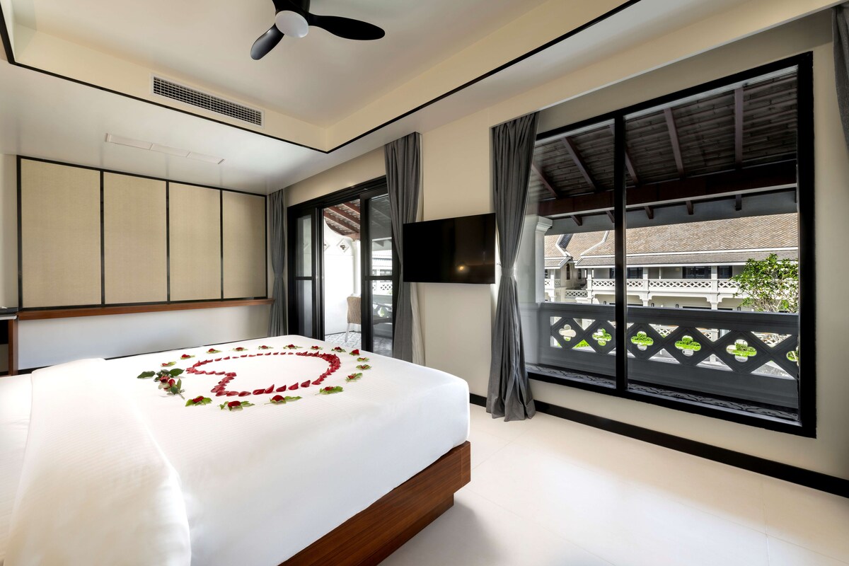 Quarto de hotel com cama grande, ventilador de teto, televisão e varanda com vista para o edifício.