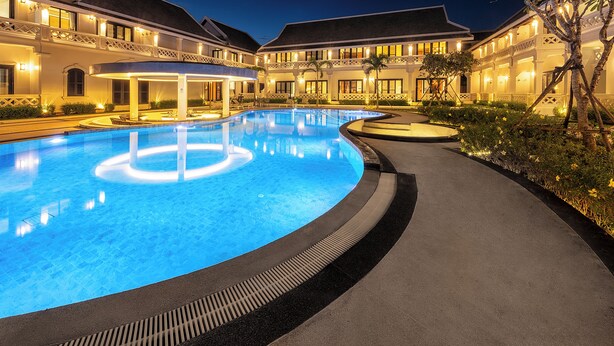Una gran piscina iluminada, rodeada por un edificio bien iluminado con varios balcones y un jardín ajardinado.