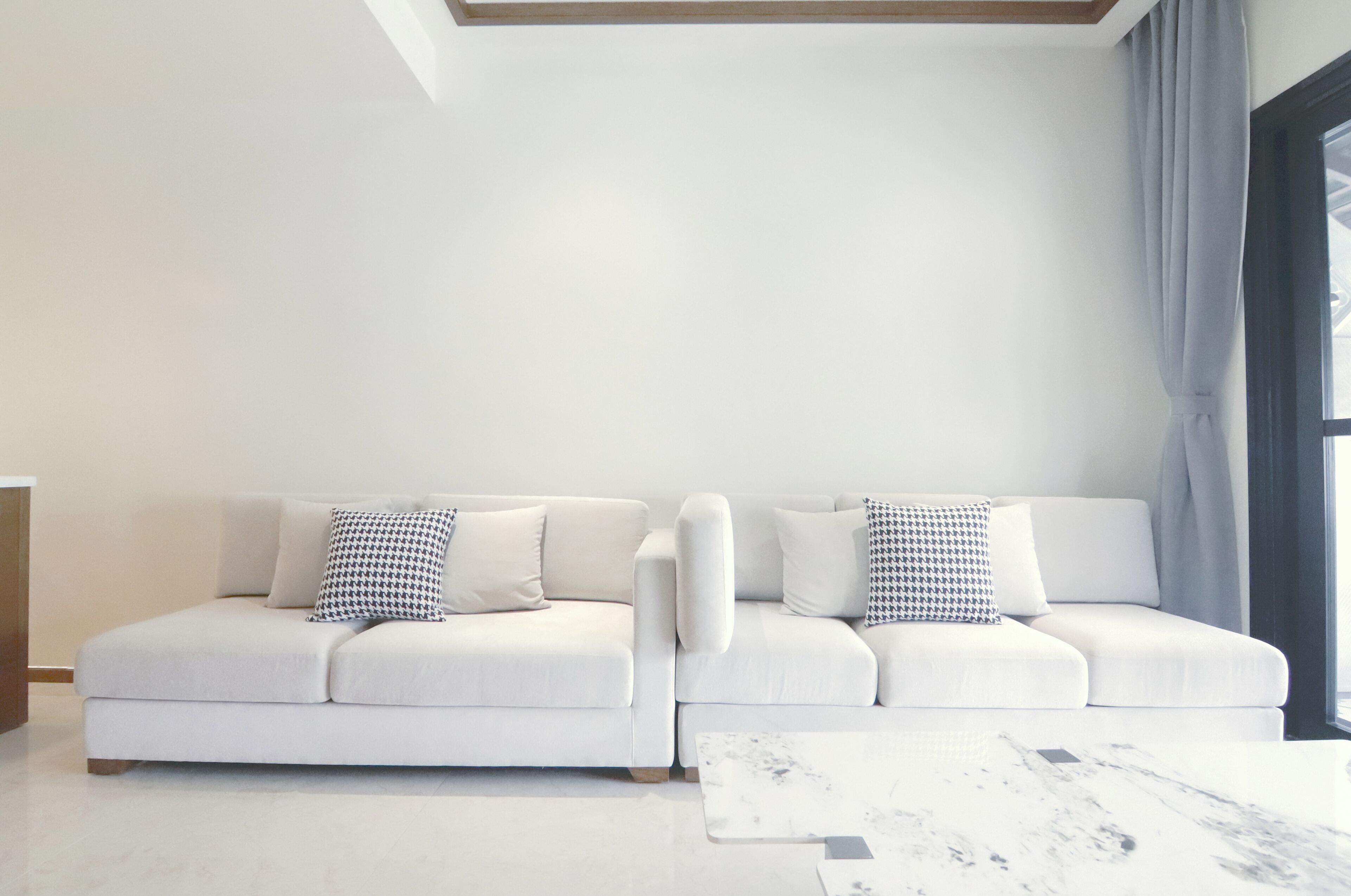 A modern living room with a white sofa set, a marble coffee table, and a large window with curtains.