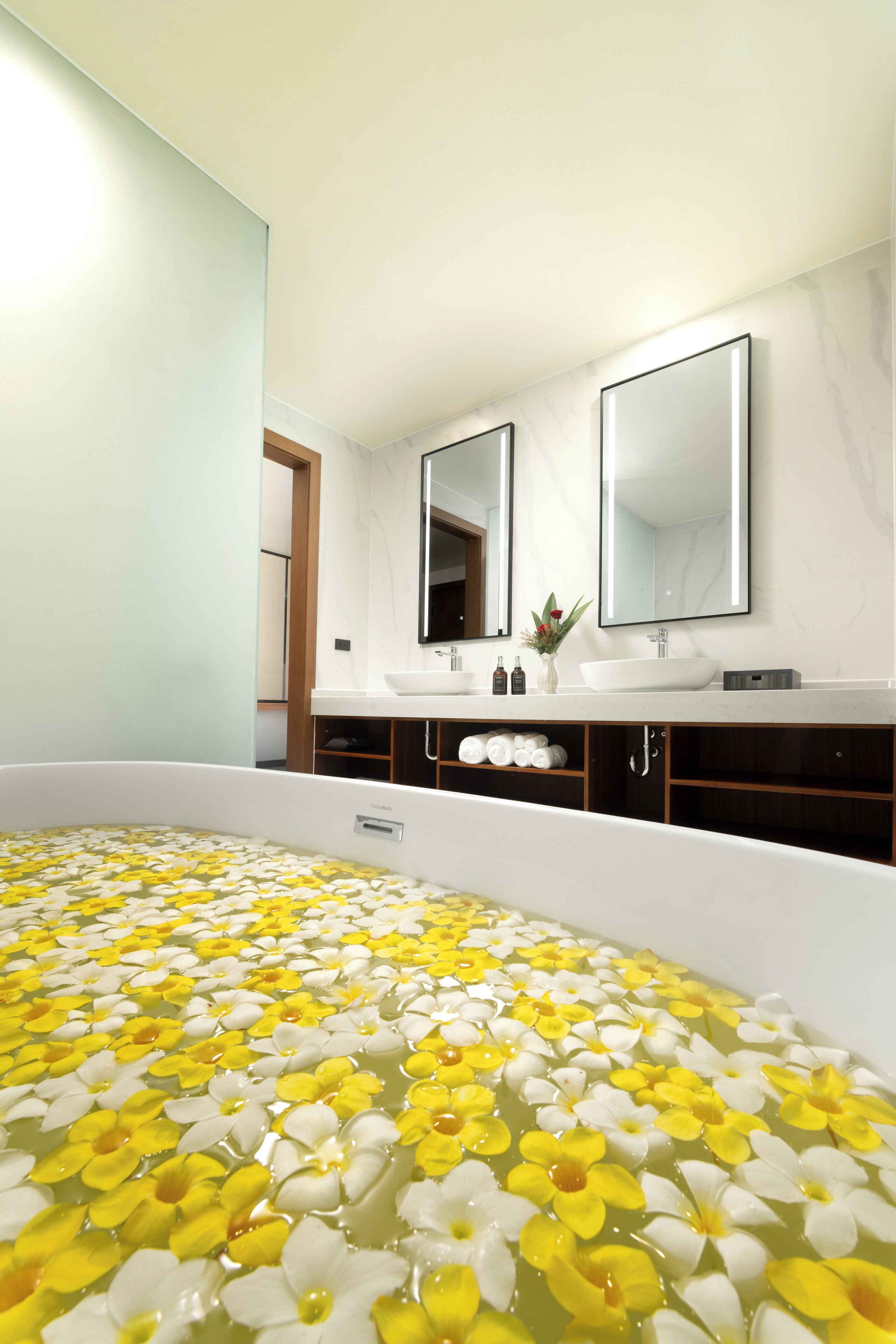 A modern bathroom with a large bathtub filled with yellow and white flowers, two mirrors, a white sink, and a wooden cabinet.