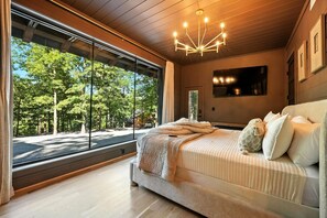 Une chambre moderne avec un grand lit, un lustre et une vue sur les arbres à travers de grandes fenêtres du sol au plafond.