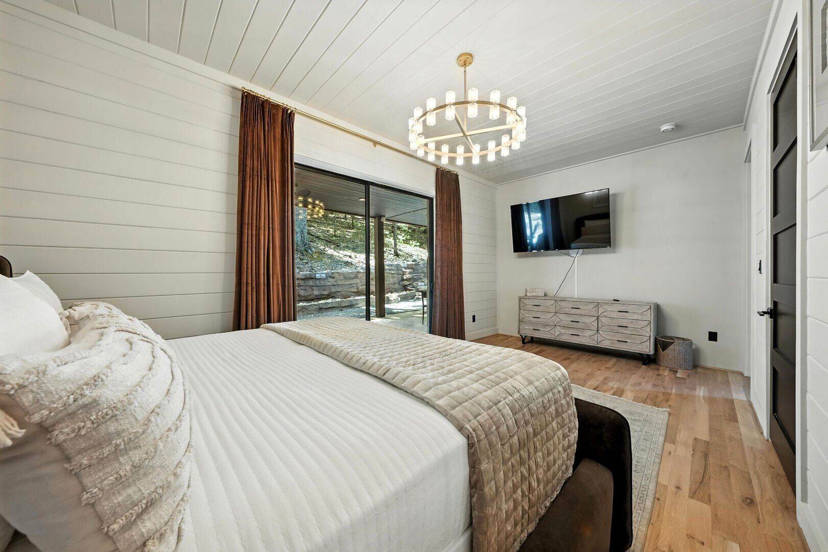 A bedroom with a large bed, a TV mounted on the wall, and a view of the outdoors through a sliding glass door.