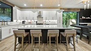 A modern kitchen with white cabinets, a large island with bar stools, pendant lighting, and a view of greenery outside. - Cave & Cascade: Rooftop Deck & Pickleball (Broken Bow)