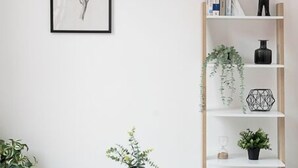 Een minimalistisch ingerichte kamer met een witte tafel, grijze stoelen, een boekenplank met boeken en decoratieve objecten, en een lijst met kunstwerk aan de muur.