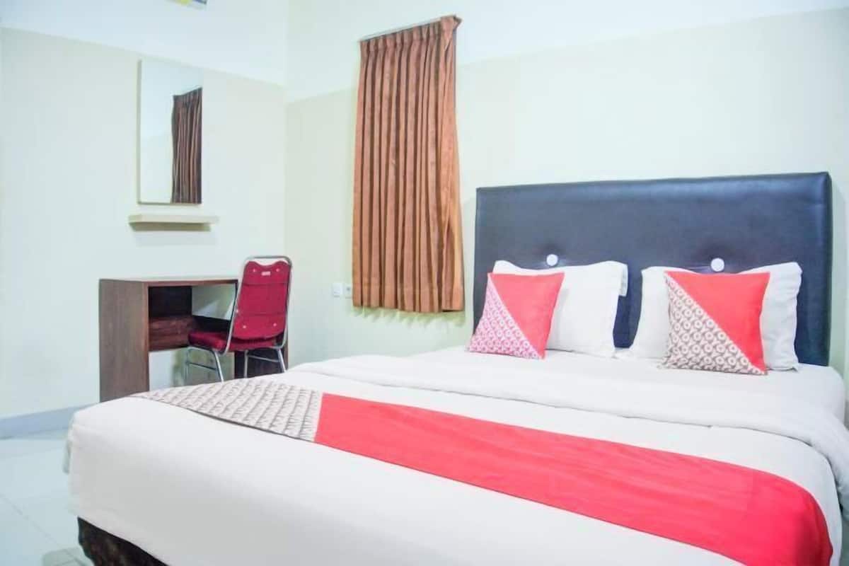 Habitación de hotel con cama, escritorio, silla, ventana con cortinas y espejo en la pared.