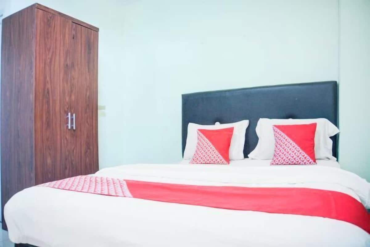 Una cama con cabecera oscura, dos almohadas, una colcha roja y blanca, y un armario de madera.