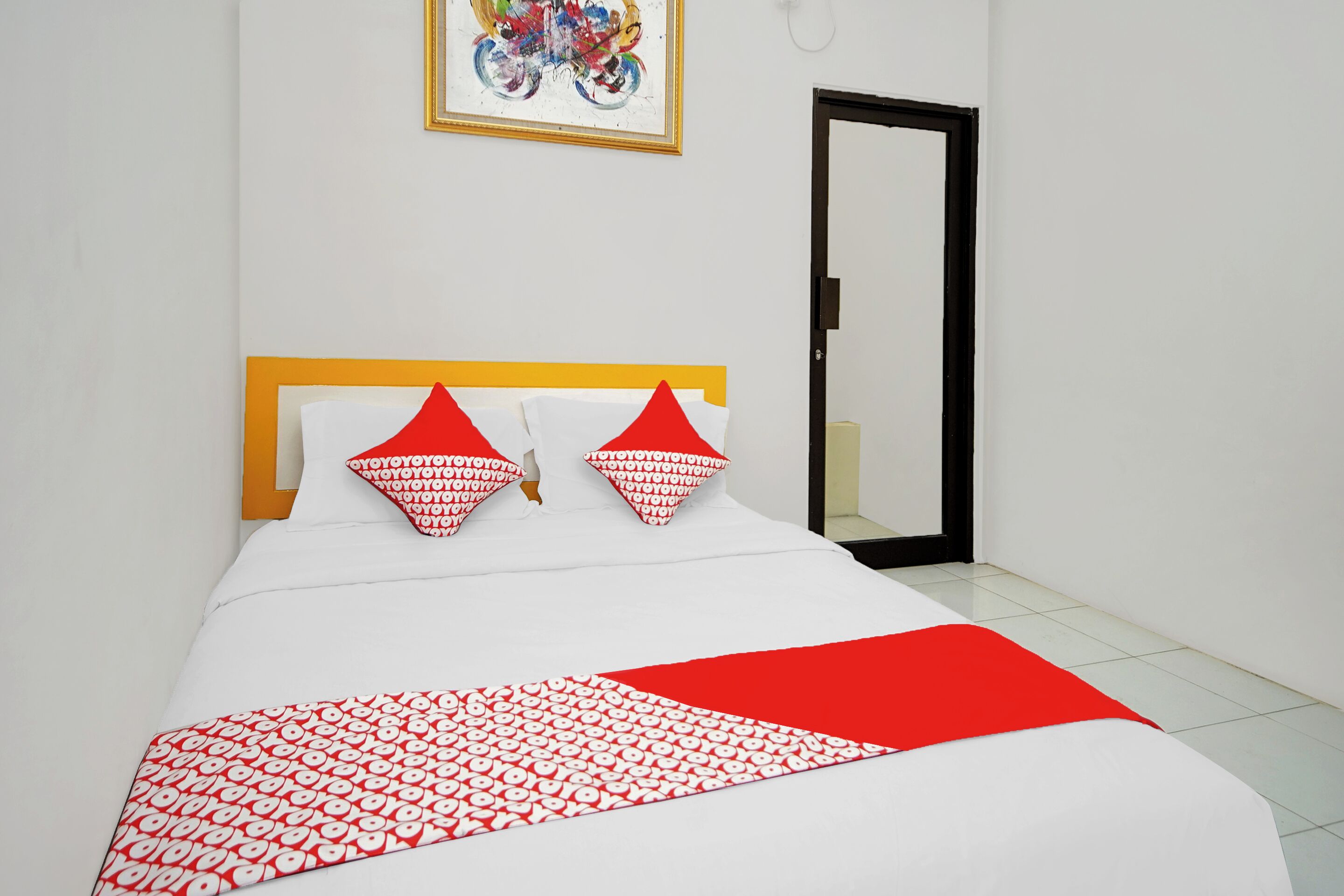 Een net opgemaakt bed met een wit en rood gebedrukte sprei, een houten hoofdbord en twee rode kussens met een wit patroon.
