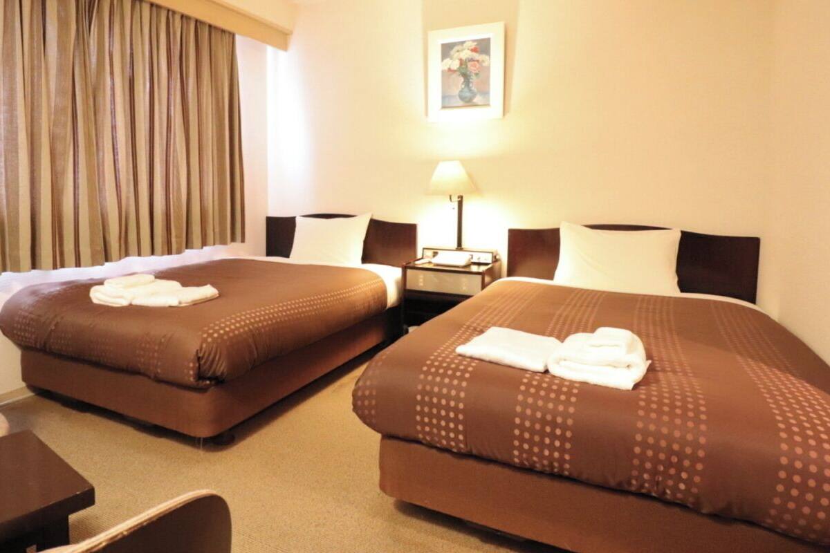 A hotel room with two beds, a chair, a small table, a lamp, and a painting on the wall.