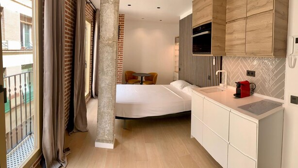 A compact hotel room with a kitchenette, a bed, and a balcony with curtains. - Exterior Studio 3rd floor (Málaga)