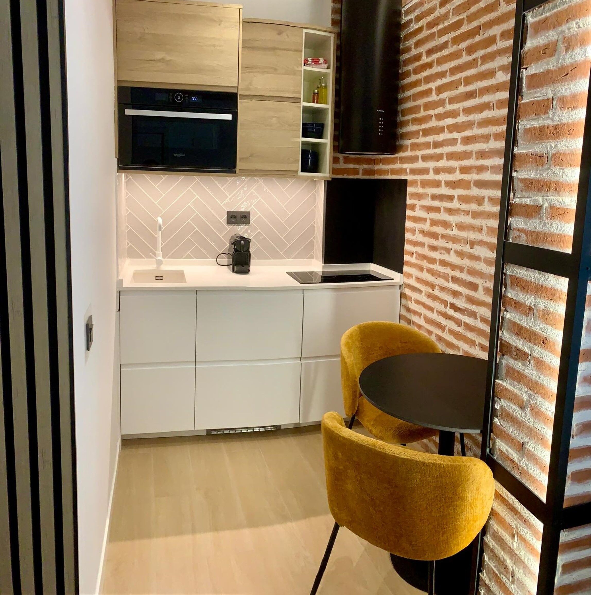 A modern kitchen with white cabinetry, a built-in oven, and a black range hood. There is a yellow chair and a small round table in the dining area.